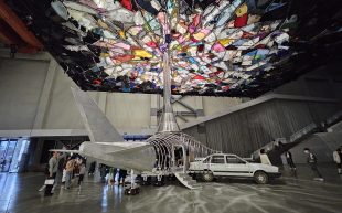 'Yin Xiuzhen: Piercing the Sky', installation view at Power Station of Art. A metal plane-like structure with a sedan attached and a needle reaching into a mosaic of clothing installation near the ceiling.