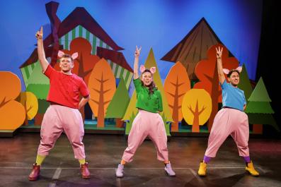 Two men and a woman dress up as three little pigs. They have pink pants on and pig ears. They are wearing a red, green and blue top respectively in 'The 3 Little Pigs.'