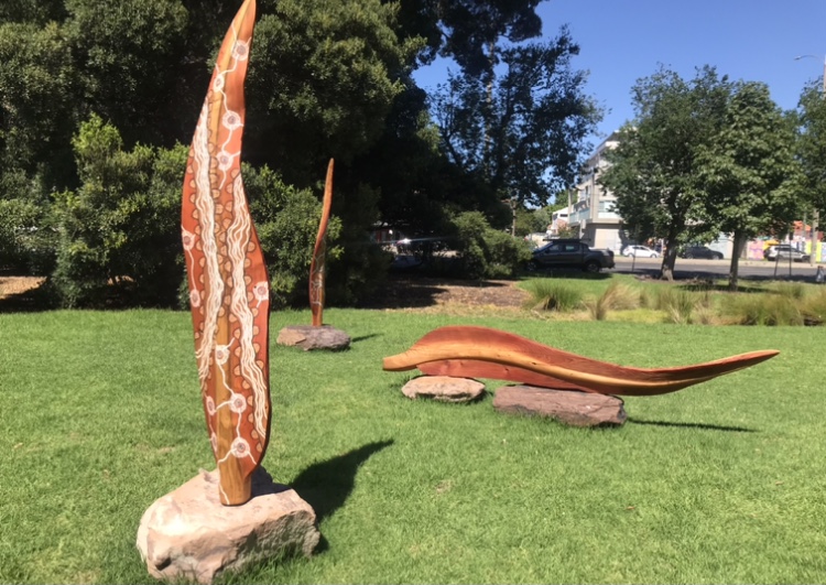 public art in the form of large leaves with Indigenous markings on them, two standing on rocks and one lying. They're all placed on grass near trees.