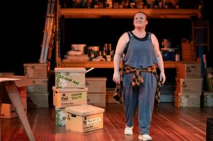 Local playwright Danielle Brame Whiting's 'Flat Pack' was supported by The Art House from concept to stage. The photo shows a scene from the play: a woman in overalls, with a flannelette shirt tied around her waist, stands on stage surrounded by boxes and stacked shelves.