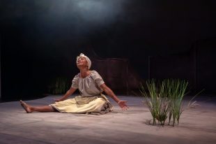 In the production of 'Ilarun' a black woman in white, with a white turban, is sitting down wit h her arms apart. On the right is a tuft of plants.