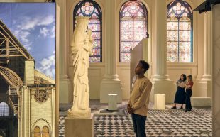 ‘Notre-Dame de Paris: The Augmented Exhibition’ at Immigration Museum. An exhibition space with large panels depicting the architecture of a church and a sculpture of Mary. A person is looking up at the sculpture while two more people behind them are looking at a screen.