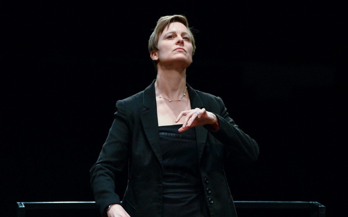 Willoughby Symphony Orchestra Associate Conductor, Dr Sarah Penicka-Smith conducting on a stage. She is a middle-aged Caucasian woman with pale skin and short blond hair, wearing a black blazer. Her left hand is raised and her head held high with a concentrated expression.