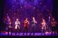 The cast of 'Love Actually? The Musical Parody' on stage. There are Christmas tress behind then and two large lit up arches with a bow in the middle.