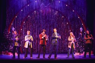 The cast of 'Love Actually? The Musical Parody' on stage. There are Christmas tress behind then and two large lit up arches with a bow in the middle.