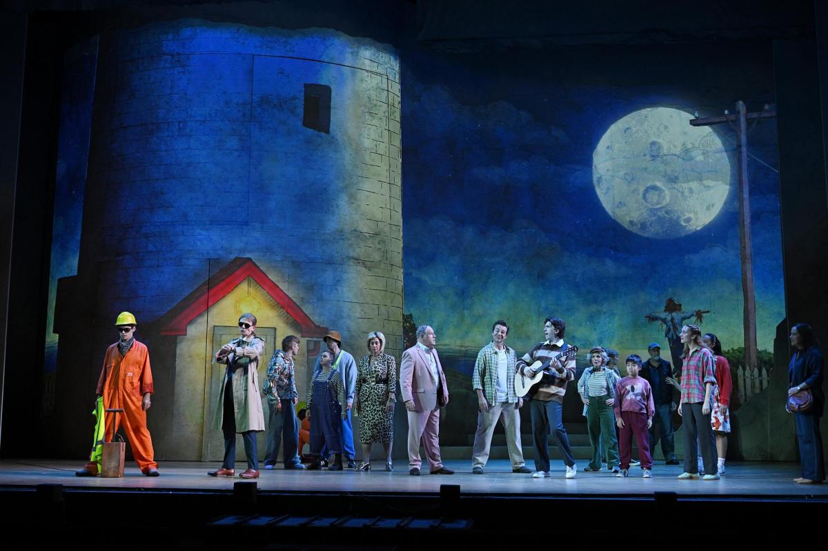 A crowd scene from "Round The Twist The Musical'. People are standing against a backdrop of a building and a moon.