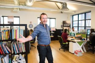 Martin Hughes, wearing a blue shirt and dark troursers, has his arms spread out in the office of publishing firm Affirm.