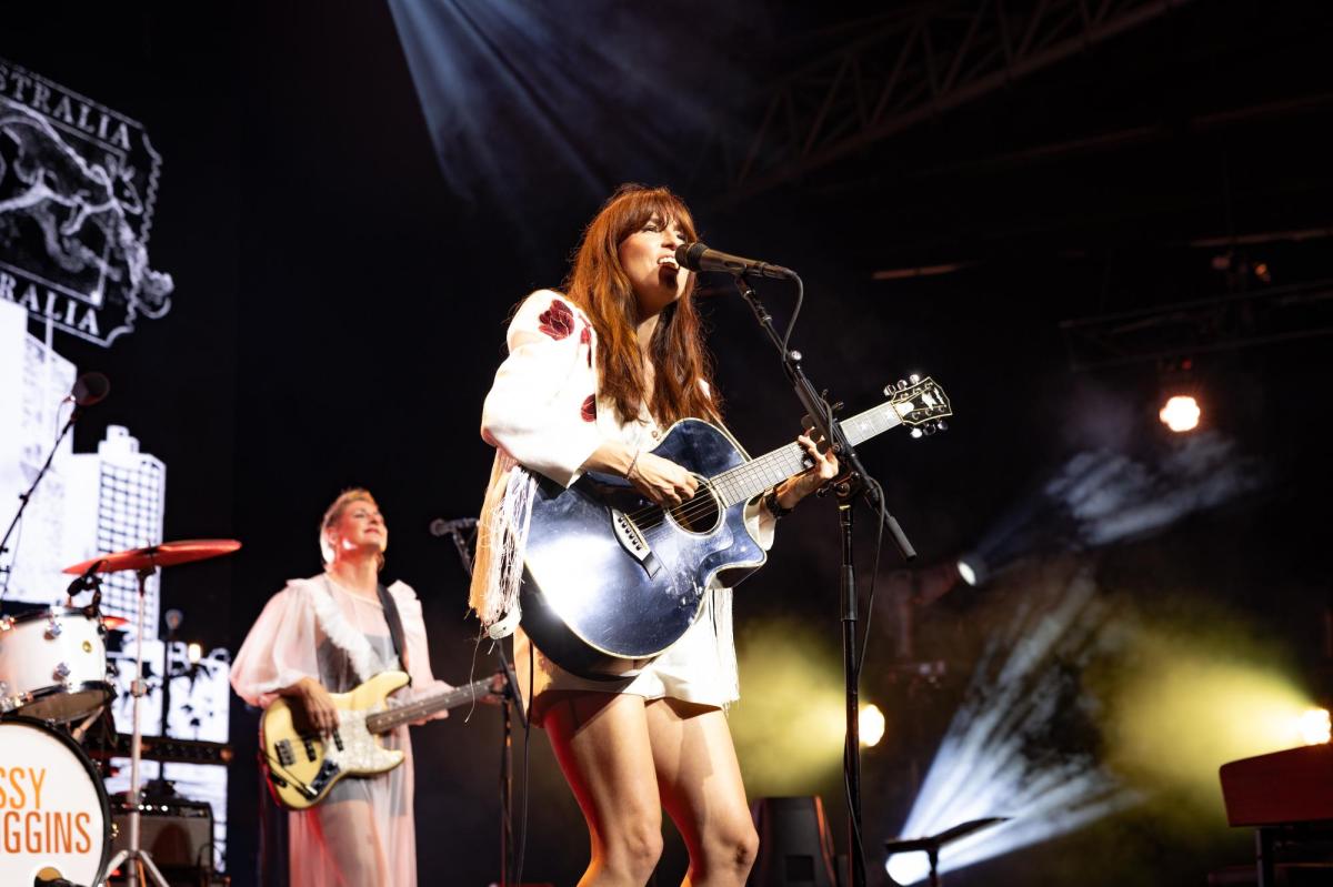 Singer Missy Higgins wearing a white pantsuit and holding a guiltar on stage. Behind her on the left is another musician playing the guitar.