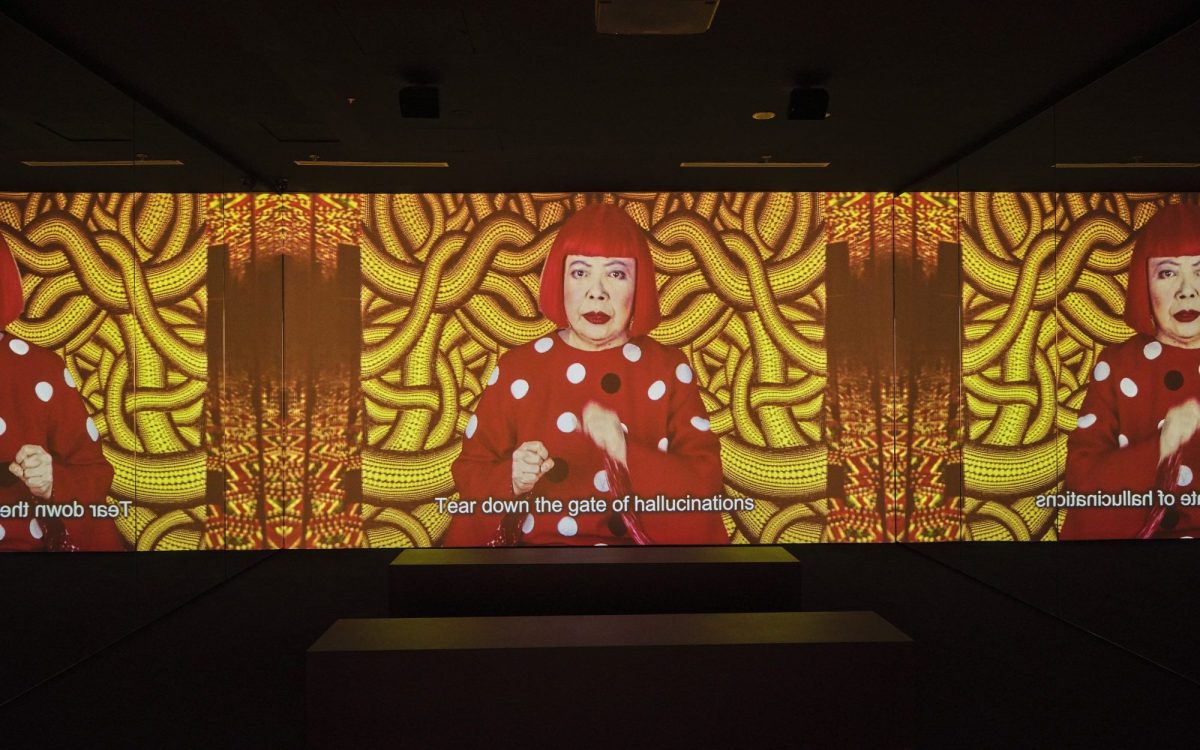 Yayoi Kusama in 'Song of a Manhattan Suicide Addict', 1978, installation view at NGV International. An old Asian woman with bright red bob and red and white polka dot top, gesturing and singing on screen. Text in the subtitle reads "Tear down the gate of hallucinations".