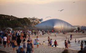 Sculpture by the Sea: a crowded beach at sunset where people are walking around a large metal sculpture in the shape of a giant wine/ goon bag which is installed by the water's edge.