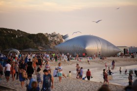 Sculpture by the Sea: a crowded beach at sunset where people are walking around a large metal sculpture in the shape of a giant wine/ goon bag which is installed by the water's edge.