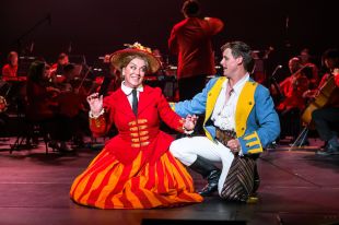 Prospero Arts and QPAC's 2024 production of Gilbert and Sullivan's 'The Pirates of Penzance – in Concert'. Two actors kneel on the stage, with the orchestra and conductor visible behind them. The woman wears a red and orange Victorian dress and straw hat; she is smiling broadly at a young man in a blue and yellow jacket, opened to reveal his white short and white trousers. He wears high black boots and a colourful scarf dangles from his waist.