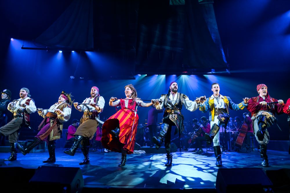 Prospero Arts and QPAC’s 2024 production of Gilbert and Sullivan’s ‘The Pirates of Penzance – in Concert’. Eight cast members dance and sing in a line across the stage, all wearing colourful piratical costumes save one man dressed as a policeman and a woman in a Victorian-era dress. Everyone is holding fencing foils and kicking their left legs into the air as they sing.