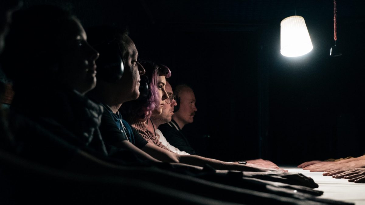 Five people in profile with headphones on. They are sitting in the dark with their hands on a table in front of them.