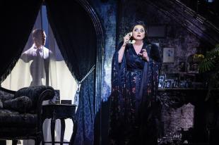 Sarah Brightman wearing a dark robe and holding onto an old-fashioned cream phone in a drawing room. On the left is a figure of man seen through the curtains.