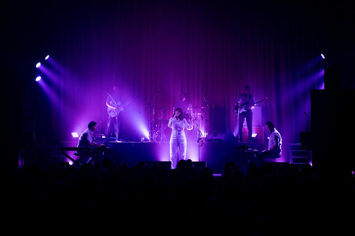 Sarah Blasko, in white, is in the centre of the stage. Five musicians surround her. The lighting is in shades of purple.