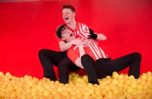 Sterling Notley and Matt Hogan in Canberra Youth Theatre's production of 'Work, But This Time Like You Mean It'. Two young men, both dressed in red and white shirts and black pants, sit in a pit of yellow balls against a red wall. One boy holds the other in his arms; both are shouting or screaming.