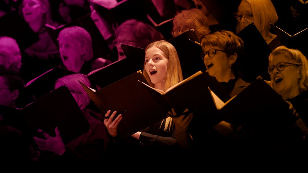 The hero image for Sydney Philharmonic Choirs' 2025 season. The photo shows the faces of 10 female choristers of various ages singing, their mouths open, and scores held in black-bound folders in their hands.