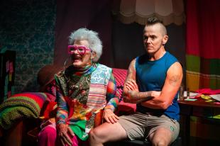 Margi Brown Ash, a old woman wearing pink glasses and colourful clothes is sitting next to Zac , a trans man in shorts and a blue tank top in 'Wanderings'.