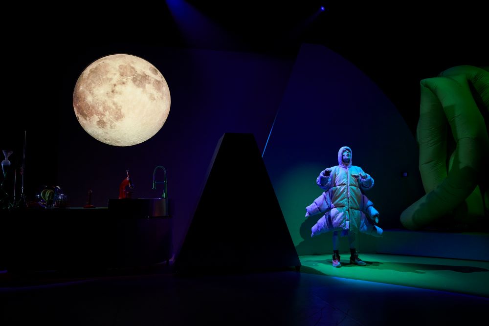 Exploring the world of tardigrades, Windmill's 'Moss Piglet' returns in 2025. An actor dressed as an eight-legged tardigrade poses on stage under a blue-green spotlight, a digitally projected moon rising behind them.