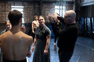 Artistic Director Yaron Lifschitz (right) in rehearsal with the Circa ensemble. A shaven-headed man in black-framed glasses and black clothes gesticulates as he explains something to a group of shirtless men and women in dark green dresses in a rehearsal studio.