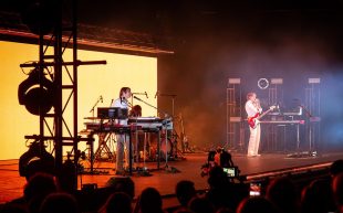 A concert scene with two musicians on stage. The one on the left is surrounded by keyboards. The one on the right has a guitar.