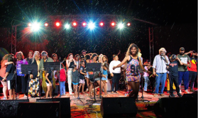 A large group of First Nations people, young and old, stand on a stage beneath spotlights and behind some music stands. At the front is a young woman with her arms out wearing multicoloured tops and shorts.