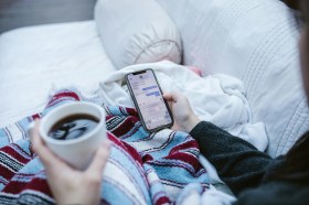 overhead detail view of person on lounge with coffee, rug and reading phone. Arts news