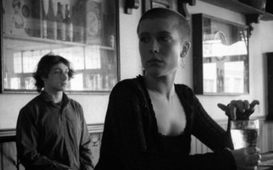 A black and white photo of Callum O'Mara, Shannon Stevens in Splinter'. They are at a bar. She is holding onto a drink at the bench on the right. He is a little distance behind her on the left.