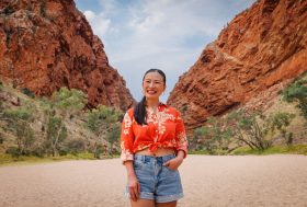 Poh Ling Yeow shown in the photo, standing in a natural landscape with two hilly terrains converging behind her back. She is a middle-aged Asian woman with a low black pony tail, wearing an orange shirt tied at the waist with denim shorts, smiling warmly.