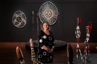 Woman with curly hair wearing black dress with white circles and surrounded by Aboriginal art. Nici Cumpston