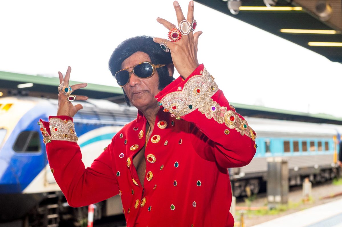 An Elvis impersonator in a red jump suit holds up his hands on the platform next to a blue train. Parkes Elvis Festival.