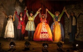 'The Lewis Trilogy' by Griffin Theatre Company, nominated for Best Mainstage Production at the 2024 Sydney Theatre Awards. Six performers dressed in colourful, elaborate stage costumes, holding hands that are raised in the air as celebration.