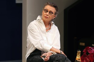 A middle aged woman with short slicked back grey hair and round black framed glasses wearing a white shirt and black pants is sitting wither legs crossed and holding a champagne flute. She has a serious expression on her face and is looking slightly off camera. Tracy Mann. Aria by David Williamson.