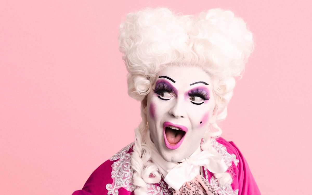 Luke Belle as Adore Händel. They are have a white beehive wig on, white makeup and are wearing a pink costume.