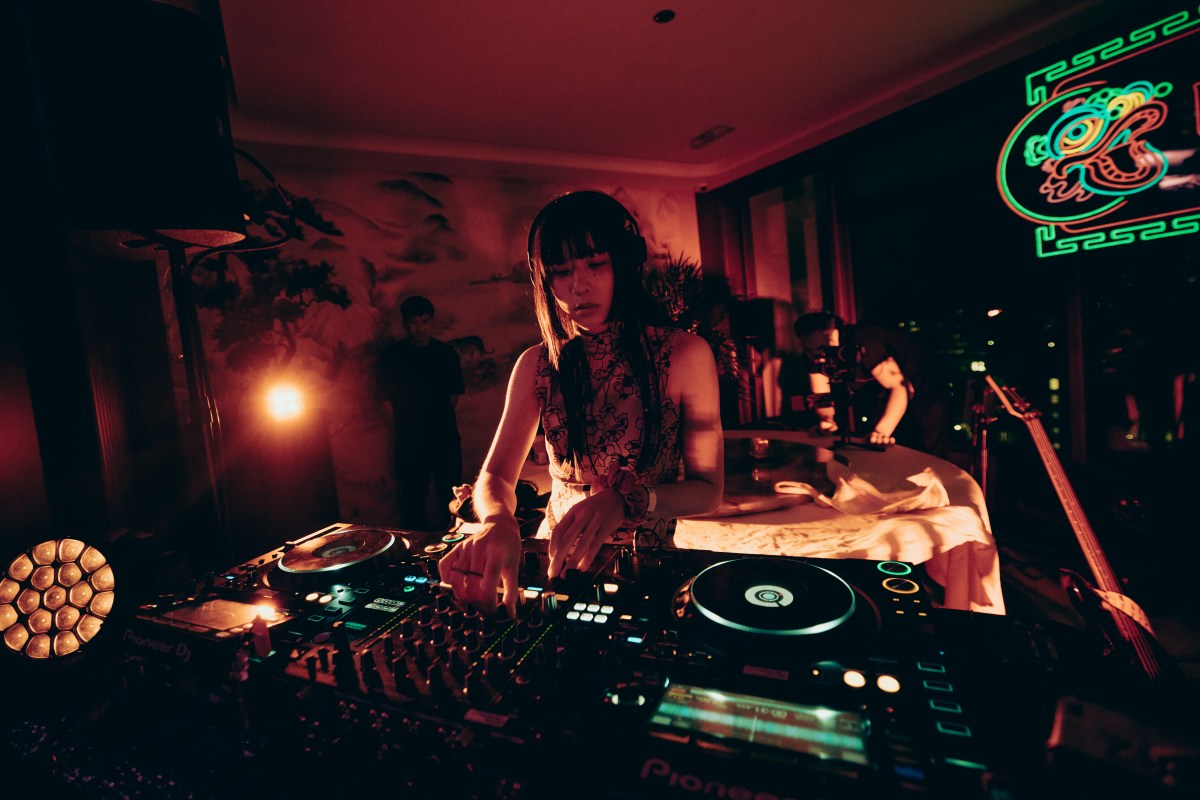 DJ Small Fry, shown in a photo behind the turntable wearing a cheongsam top with long black hair and bangs, inside a small room with low, warm coloured light.