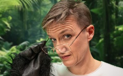 Kate Smurthwaite, a woman with short brown hair and glasses is wearing a white tee shirt and a black furry ape glove.