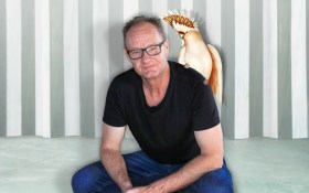 Rob Carlton seated with a cockatoo on his shoulder. He is a Caucasian man with balding hair and glasses, wearing a black tshirt and jeans.