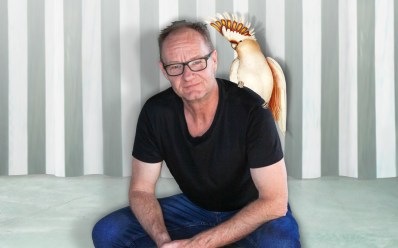 Rob Carlton seated with a cockatoo on his shoulder. He is a Caucasian man with balding hair and glasses, wearing a black tshirt and jeans.