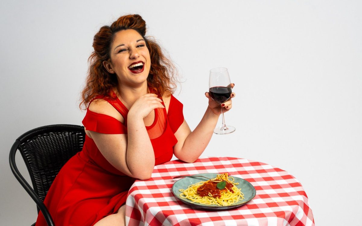 Ileana Pipitone, a woman in a red dress holding a glass of wine. She's seated at a red and white checked tablecloth with a bowl of spaghetti on it.