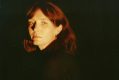 Black backdrop for a head and shoulders shot of a young woman with red hair, fringed and parted in the middle. She is three-quarters on, looking to the right of the photographer and not smiling. Sarah Blasko.