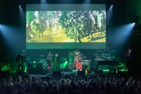 A concert scene with a large screen in the background showing foliage.