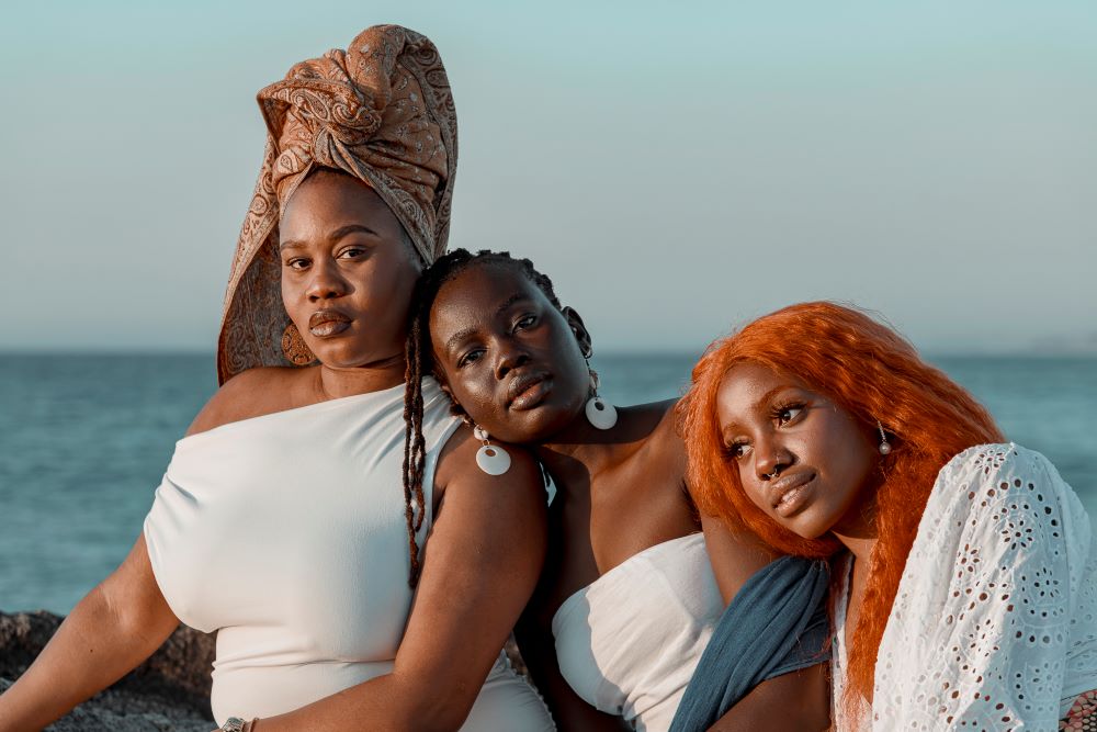 A publicity image for 'Our Stories, Our Motherland' at Fringe World 2025. The photo depicts three women of African heritage leaning on and physically supported by one another. The woman on the left wears a white dress and a colourful head scarf and is staring directly at the camera; the woman in the centre has dangling white, circular earrings  and dreadlocks, and leans her her on the shoulder of the woman to her left; she is also staring at the canera. The third woman has red-dyed hair and wears a white dress; she leans on the shoulder of the woman to her left in tirn. 