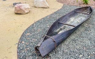 Bronze canoe stolen as part of public artwork, shown prior to its theft. A canoe made out of bronze laying on the ground in an outdoor setting next to some rocks.