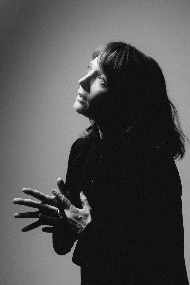 Black and white shot of top half of Caucasian 30-something woman, side on, wearing black, looking up and holding her hands in front of her with outstretched fingers.