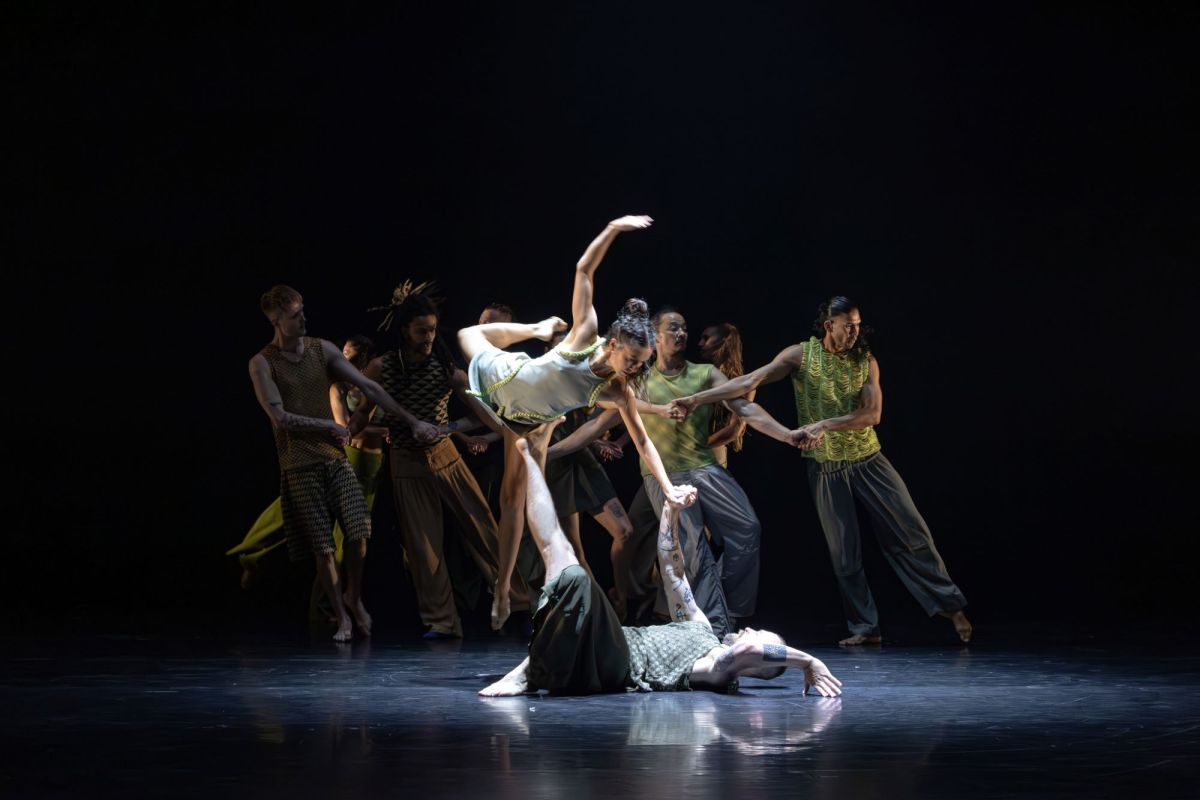 Stephanie Lake Company’s ‘The Chronicles’ is sure to be a highlight of Sydney Festival 2025 and indeed, the 2025 summer festival season generally. The photo shows five dancers of diverse cultural backgrounds holding hands and collectively leaning to the right, while in the fourground, and much more nrightly lit, a male-presenting dancer lies on their back on the stage, holding up a female-presenting dancer with one hand and one foot. Balance upon his raised extremities, the female-presenting dancer has her right leg and right arm raised in a snapshot of dynamic movement.