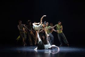 Stephanie Lake Company’s ‘The Chronicles’ is sure to be a highlight of Sydney Festival 2025 and indeed, the 2025 summer festival season generally. The photo shows five dancers of diverse cultural backgrounds holding hands and collectively leaning to the right, while in the fourground, and much more nrightly lit, a male-presenting dancer lies on their back on the stage, holding up a female-presenting dancer with one hand and one foot. Balance upon his raised extremities, the female-presenting dancer has her right leg and right arm raised in a snapshot of dynamic movement.