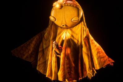 A woman is peeping through the folds of a large, oversized puppet. The light on them both is golden in a production of 'Hometown.'