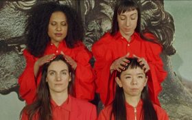 Sara Cwynar, ‘Red Film’, 2018, to be presented by Cooper Cole at the Melbourne Art Fair 2025. The photo shows four female-presenting figures dressed in red collared shirts. Two are sitting while two stand behind them, with their hands on the heads of those seated.