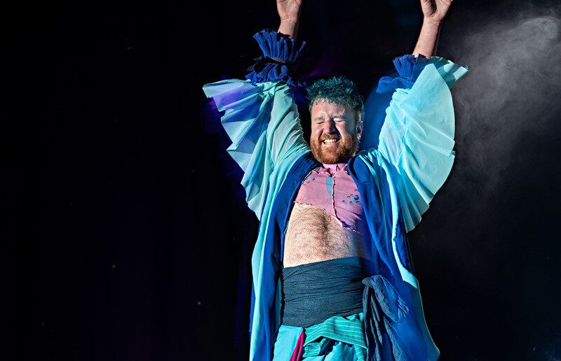 A man, Timothy Christopher Ryan, with a bare midriff wearing an aqua robe and pants raising his arms above his head.